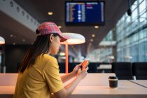 Side view of asian woman in cap surfing mobile phone and drinking coffee from disposable cap at table in airport — Stock Photo