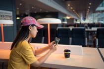 Азіатська жінка в кашкеті серфінг мобільного телефону і випивання кави з одноразового кашкета за столом в аеропорту. — стокове фото