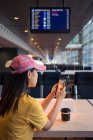Rückansicht einer Frau mit Schirmmütze, die am Tisch im Flughafen Handy surft und Kaffee aus einer Einwegkappe trinkt — Stockfoto