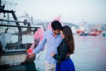 Loving couple embracing on pier with city harbor on background standing with pink smoking bomb — Stock Photo