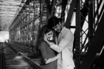Homem amoroso e mulher macia em vestido azul abraçando na ferrovia sob construção de ponte de metal — Fotografia de Stock