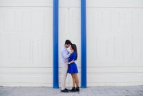 Vista lateral de homem amoroso e mulher em roupas elegantes beijando no fundo da parede branca com colunas azuis brilhantes — Fotografia de Stock