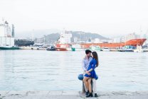 Side view of charming woman in blue dress in embrace with handsome man bonding on pier of city port with ship on background — Stock Photo