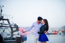Любящая пара обнимается на пирсе с городской гаванью на заднем плане стоя с розовой бомбой для курения — стоковое фото