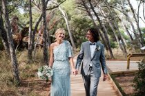 Jeune couple nouvellement marié en tenue de mariage marche, tenant la main sur le chemin parmi la belle forêt verte avec de grands arbres — Photo de stock