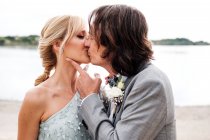 Piacevole giovane sposo in abito da sposa abbracciare e baciare sposa dai capelli biondi in abito elegante dietro a riva di sabbia vuota — Foto stock