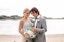 Jovem noivo satisfeito em terno de casamento orgulhosamente olhando para bela noiva de cabelos loiros em vestido elegante atrás na costa — Fotografia de Stock