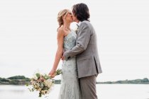 Zufriedener junger Bräutigam im Hochzeitsanzug umarmt und küsst blondhaarige Braut im stilvollen Kleid hinter der Küste — Stockfoto