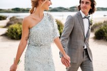 Pleased young groom in wedding suit proudly looking at beautiful blonde haired bride in stylish dress behind at seashore — Stock Photo