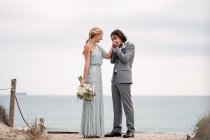 Удовлетворенный молодой жених в свадебном костюме целует руку блондинки невесты в стильном платье позади на пустом песчаном побережье — стоковое фото