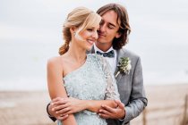 Задоволений молодий наречений у весільному костюмі обіймає блондинку волохату наречену в стильній сукні позаду порожнього піщаного узбережжя — стокове фото