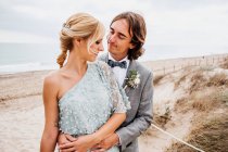 Pleased young groom in wedding suit hugging blonde haired bride in stylish dress behind at empty sandy seashore — Stock Photo