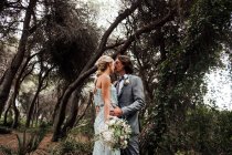 Giovane coppia di sposi in abito da sposa in piedi e abbracciandosi sul sentiero tra la bella foresta verde con grandi alberi — Foto stock