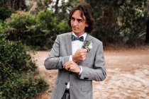 Junger Bräutigam in schickem grauen Hochzeitsanzug unterwegs auf leerer Straße mit grünen Bäumen an den Seiten — Stockfoto