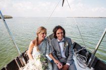 Удовлетворенные супруги в свадебной одежде отдыхают на лодке с морем на заднем плане — стоковое фото