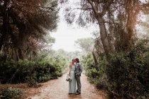 Молодая молодожёны в свадебной одежде стоят и обнимаются на дорожке среди красивых зеленых лесов с большими деревьями — стоковое фото