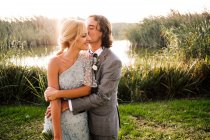 Стильна нещодавно одружена пара у весільних костюмах, яка обіймає і цілує зеленими рослинами і просто в сонячний день — стокове фото
