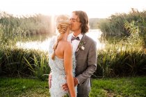 Casal recém-casado elegante em ternos casados abraçando e beijando com plantas verdes e mera no dia ensolarado — Fotografia de Stock
