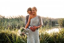 Casal recém-casado elegante em ternos casados abraçando e beijando com plantas verdes e mera no dia ensolarado — Fotografia de Stock