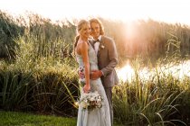 Elegante pareja de recién casados en trajes de novia abrazando y besándose con plantas verdes y mera en el día soleado - foto de stock
