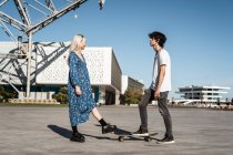 Trendy blonde Frau in übergroßen blauen Kleid und schwarzen Plateauschuhen gestoppt Skateboard und Blick auf selbstbewussten erwachsenen Mann — Stockfoto