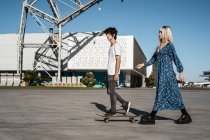 Jovem casal amante da moda andando com skate na praça contra o céu azul e edifícios modernos borrados — Fotografia de Stock