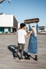 Visão traseira do jovem casal da moda em pé, enquanto a mulher levanta um skate no quadrado contra o céu azul — Fotografia de Stock