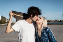 Atractiva pareja joven y elegante abrazándose y besándose en la calle mientras el hombre de camisa blanca sostiene el monopatín - foto de stock