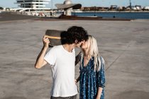 Atraente jovem estilista casal abraçando e beijando uns aos outros na rua, enquanto o homem de camisa branca segurando skate — Fotografia de Stock