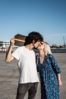 Atractiva pareja joven estilista abrazándose y besándose en la calle mientras el hombre con camisa blanca sostiene el monopatín - foto de stock