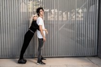 Seitenansicht des trendigen Paares, das sich gegenseitig stützt, während sich die Frau auf dem Rücken ihres Freundes gegen die gestreifte Wand des modernen Gebäudes lehnt — Stockfoto