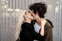 Side view of woman biting for tongue and touching neck of young curly dark haired man while standing and kissing — Stock Photo