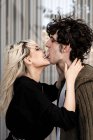Mulher loira adulto mordendo para a língua e tocando o pescoço do jovem encaracolado homem de cabelos escuros enquanto está de pé e beijando — Fotografia de Stock