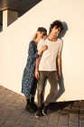 Молода стильна пара обіймає один одного, насолоджуючись і споглядаючи сонячний день, спираючись на білу стіну — стокове фото