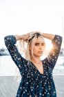 Gorgeous pensive blond female looking at camera with curiosity while standing and touching hair on street during sunset — Stock Photo
