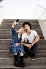Sonriente mujer adulta de moda y hombre en ropa casual tomando selfie junto con el teléfono inteligente mientras está sentado en escaleras de madera - foto de stock