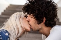 Side view of adult curly dark haired man with closed eyes kissing blond woman against blurred stairs in daylight — Stock Photo