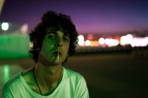 Beau gars en vêtements décontractés avec cigarette dans la bouche regardant avec intérêt la caméra sur fond flou de la rue — Photo de stock