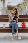 Donna in camicia a righe e jeans seduta sulla panchina di fronte al mare con tazza di caffè usa e getta e navigazione internet su tablet — Foto stock