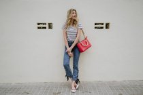 Женщина с красной сумкой в полосатой рубашке и джинсах стоит со сложенными руками и смотрит прочь с застенчивой улыбкой — стоковое фото