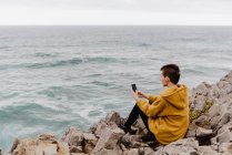 Visão traseira da mulher de cabelos curtos em camisola amarela sentada na costa rochosa e tomando selfie no telefone móvel — Fotografia de Stock