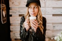 Langhaarige trendige Frau hält ein Glas leckeren schäumenden Kaffees in der Hand und leckt Lippen, die das Vergnügen des Geschmacks im Café vorwegnehmen — Stockfoto