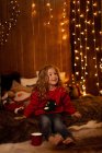 Adorable little girl with red mug sitting in room full of Christmas decoration and lights — Stock Photo