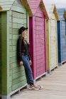 Mulher na moda em boné preto e jaqueta de couro encostada na parede de cabines de praia de madeira — Fotografia de Stock