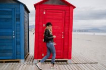 Donna alla moda in berretto nero e giacca di pelle sorridente luminosamente alla fotocamera mangiare una mela rossa mentre si cammina davanti a una spiaggia di legno cabine — Foto stock