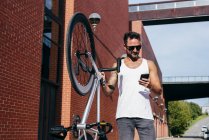 Guapo ciclista masculino en ropa deportiva y gafas de sol con teléfono inteligente mientras está de pie con la bicicleta junto a la pared de ladrillo rojo - foto de stock