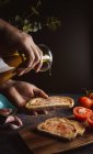 Cuoco irriconoscibile versare olio su pezzi di pane fresco con salsa durante la preparazione di toast su sfondo nero — Foto stock
