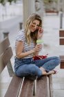 Весела випадкова жінка з чашкою кави, що сидить на міській лавці на березі моря в літній день, дивлячись на камеру — стокове фото