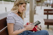 Mujer en camisa a rayas y jeans sentado en el banco de la calle en primera línea de mar con taza de café desechable y navegar por Internet en la tableta - foto de stock