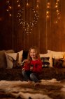 Чарівна маленька дівчинка тримає червоний кухоль, сидячи в кімнаті, повній різдвяних прикрас і вогнів — стокове фото